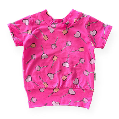 Handmade baby and toddler short-sleeve top featuring a vibrant pink lollipop and heart design. Soft, cozy, and perfect for pairing with leggings or pants for a playful yet comfortable look. Available in sizes preemie to 4T.